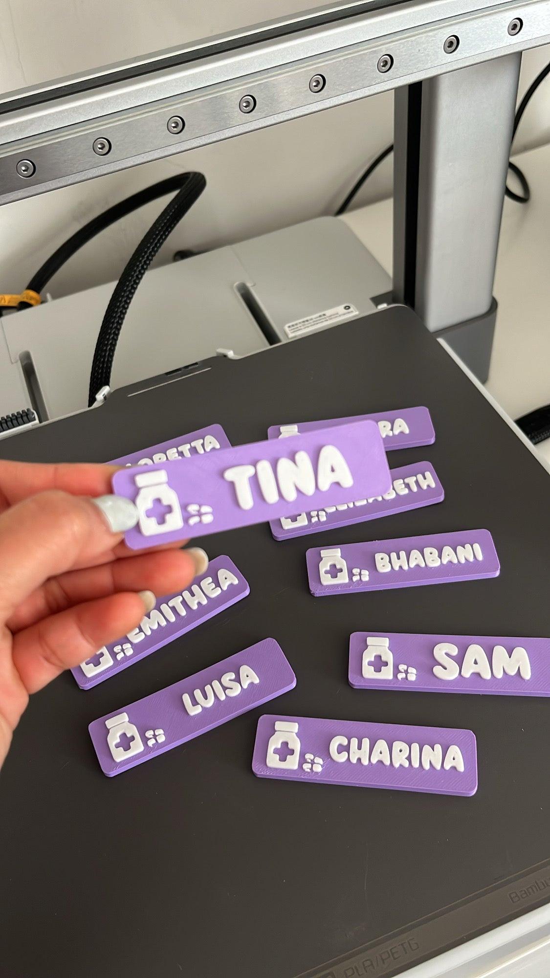 Hand holding purple name tags with names printed on them, in front of a 3D printer.
